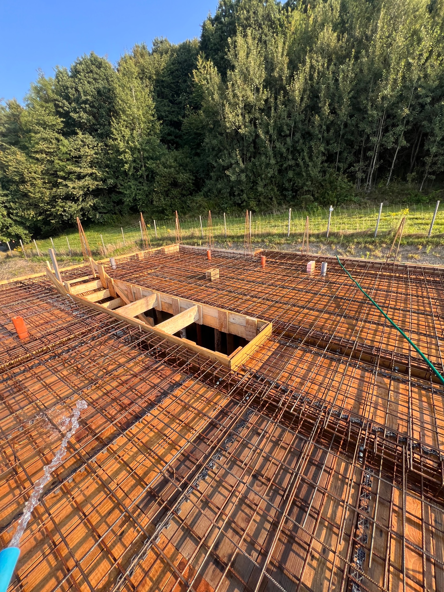 Zbrojenie fundamentów domu jednorodzinnego z drewnianym szalunkiem i wystającymi prętami. Widok z góry na przygotowany teren budowy w słoneczny dzień.