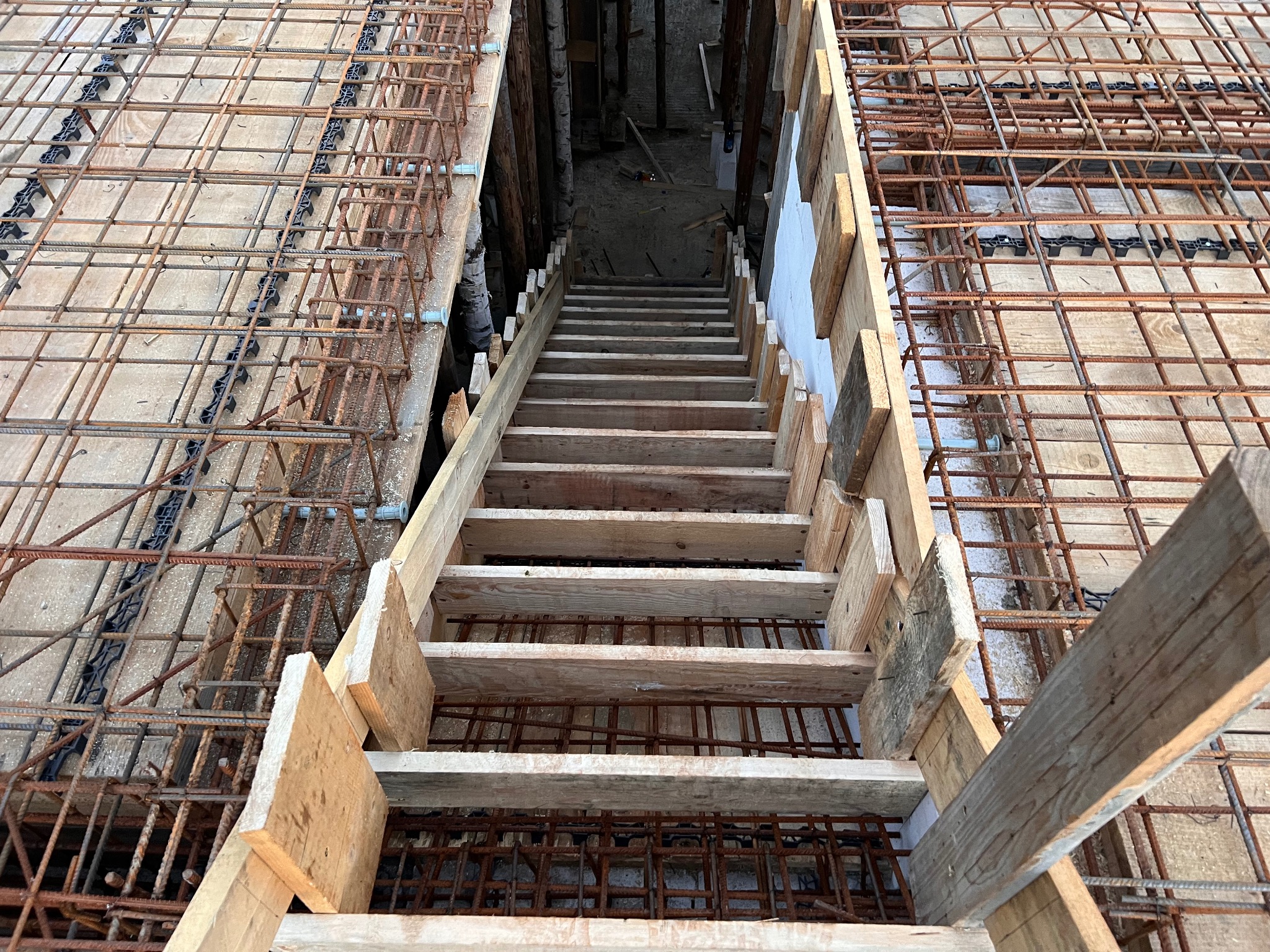 Zbrojenie stropu z drewnianym zejściem do piwnicy. Widok z góry na siatkę zbrojeniową i deskowanie. Etap prac budowlanych z użyciem stali i drewna.