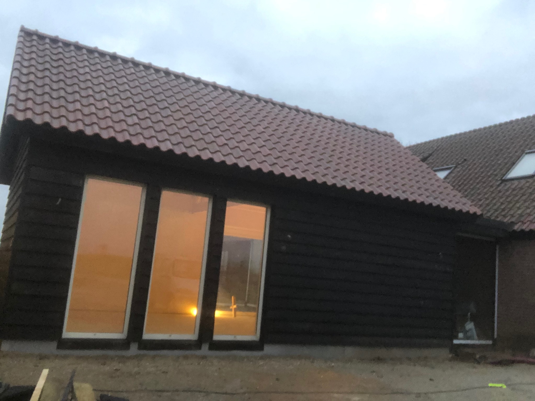 Nowoczesny budynek z ciemną drewnianą elewacją i trzema wysokimi oknami, w których odbija się ciepłe światło zachodzącego słońca, widoczny fragment dachu pokryty brązową dachówką.