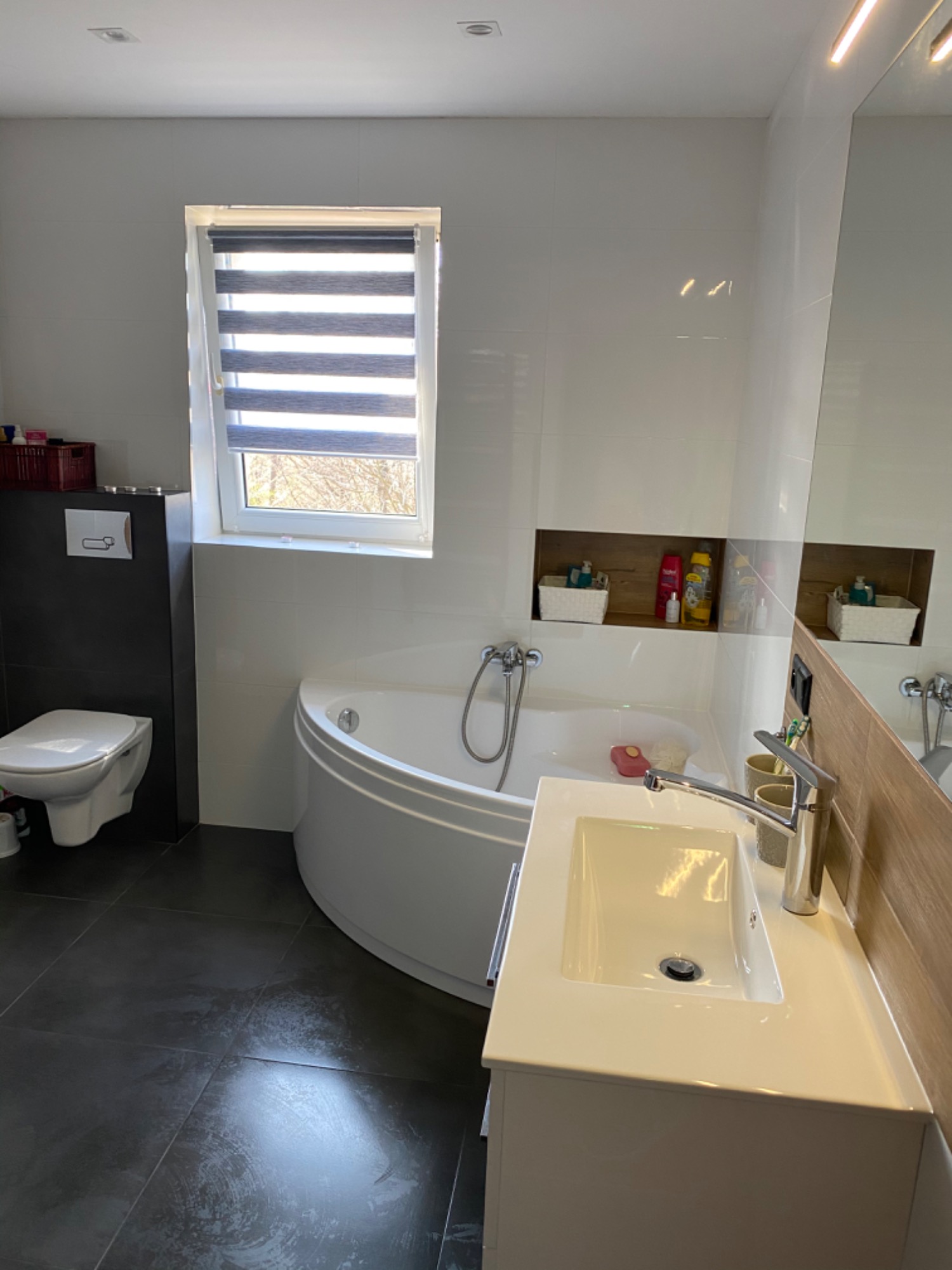 Nowoczesna łazienka z narożną wanną, umywalką nablatową, ciemną podłogą i oknem z roletami dzień-noc.