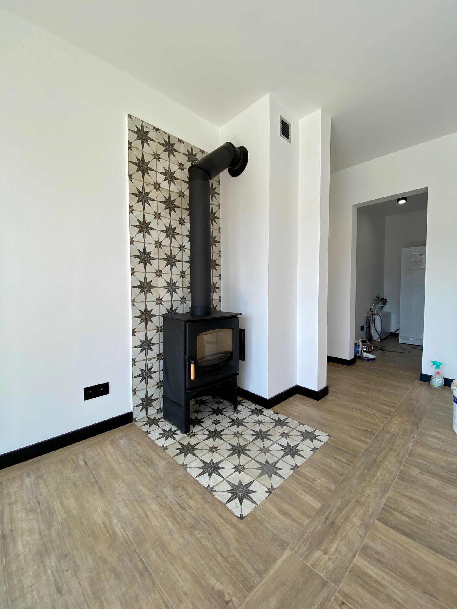 Czarny piec wolnostojący z rurą dymową na tle ściany wyłożonej dekoracyjnymi płytkami w gwiazdy, z podłogą imitującą drewno i białymi ścianami z czarnymi listwami przypodłogowymi. W tle widoczne...