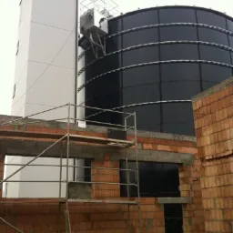 Wznoszenie budynku z cegły ceramicznej, widoczne rusztowanie i fragment konstrukcji zbiornika przemysłowego z metalu.