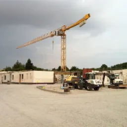 Budowa parterowego budynku z białych bloczków, widoczne maszyny budowlane i wysoki żuraw pod pochmurnym niebem.