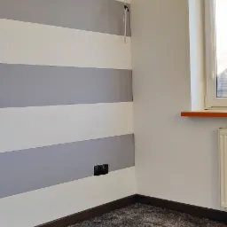 Pomalowany pokój z szarymi pasami na ścianie, wykończony listwami przypodłogowymi, z szarym dywanem na podłodze, widoczne gniazdka elektryczne i karnisz nad oknem.