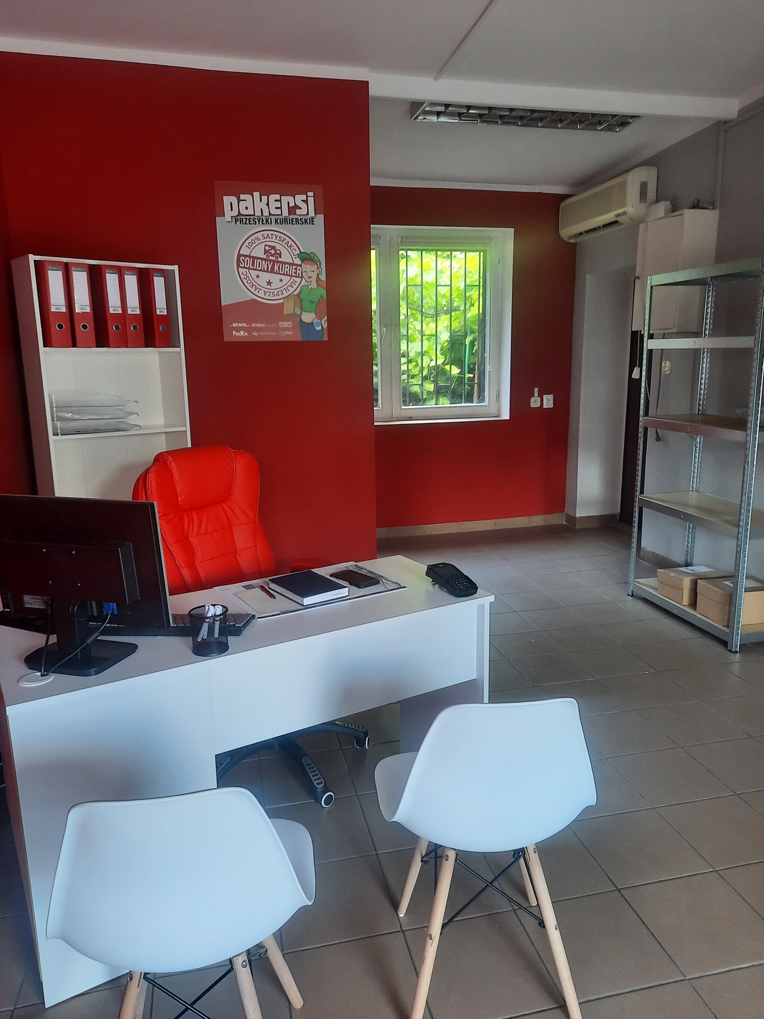 Biuro firmy kurierskiej z czerwonymi ścianami, białym biurkiem, czerwonym fotelem biurowym, regałem z segregatorami i plakatem reklamowym 'Pakersi - Przesyłki Kurierskie'.