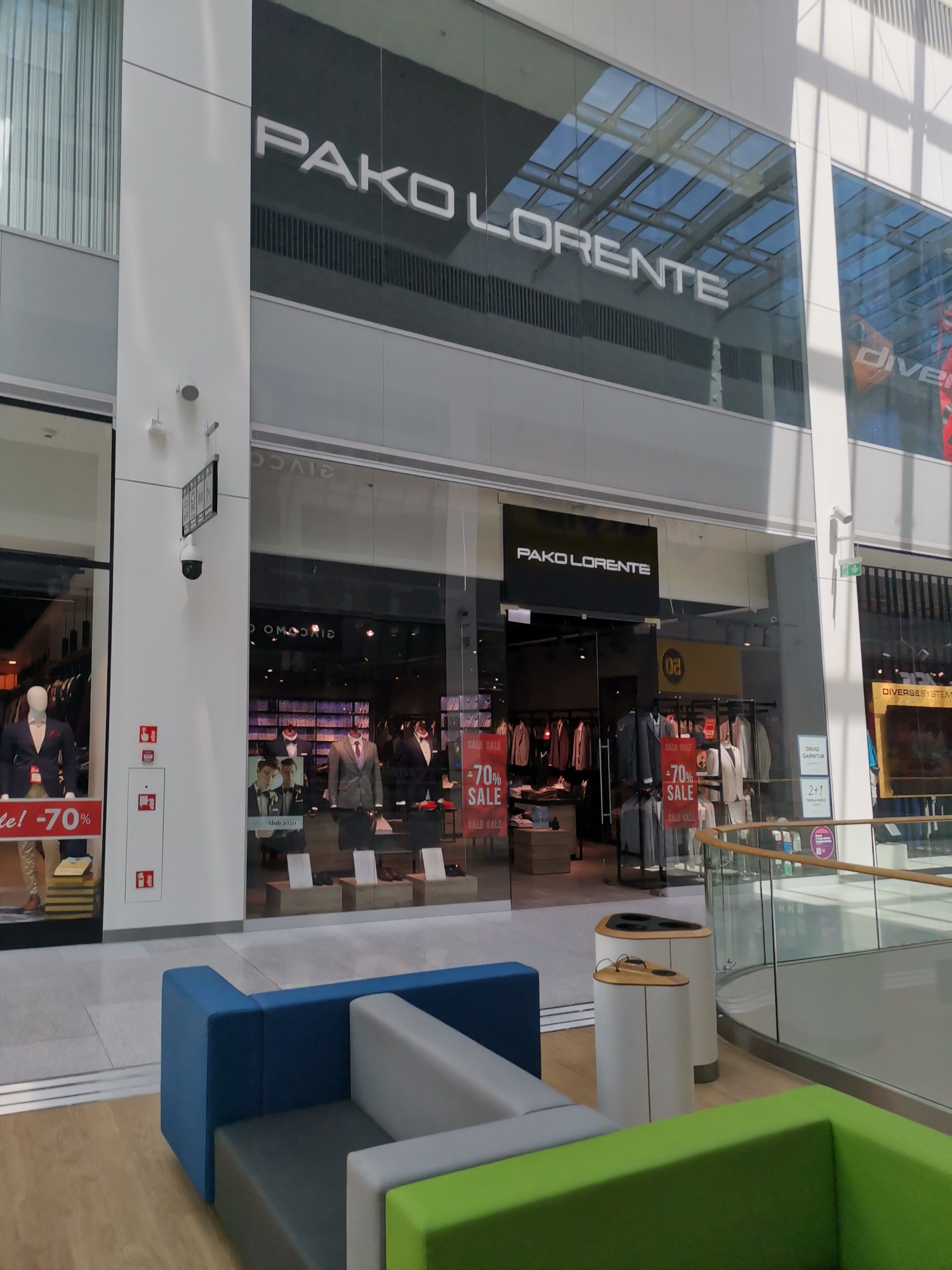 Elewacja przeszklona z widokiem na wnętrze sklepu odzieżowego Pako Lorente w galerii handlowej, widoczny napis z nazwą sklepu nad wejściem i manekiny w garniturach za szybą.