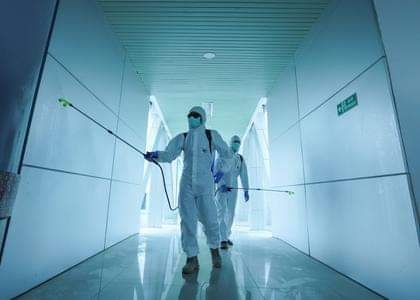 Dwóch pracowników w kombinezonach ochronnych dezynfekuje korytarz za pomocą opryskiwaczy ciśnieniowych, widoczny znak ewakuacyjny.