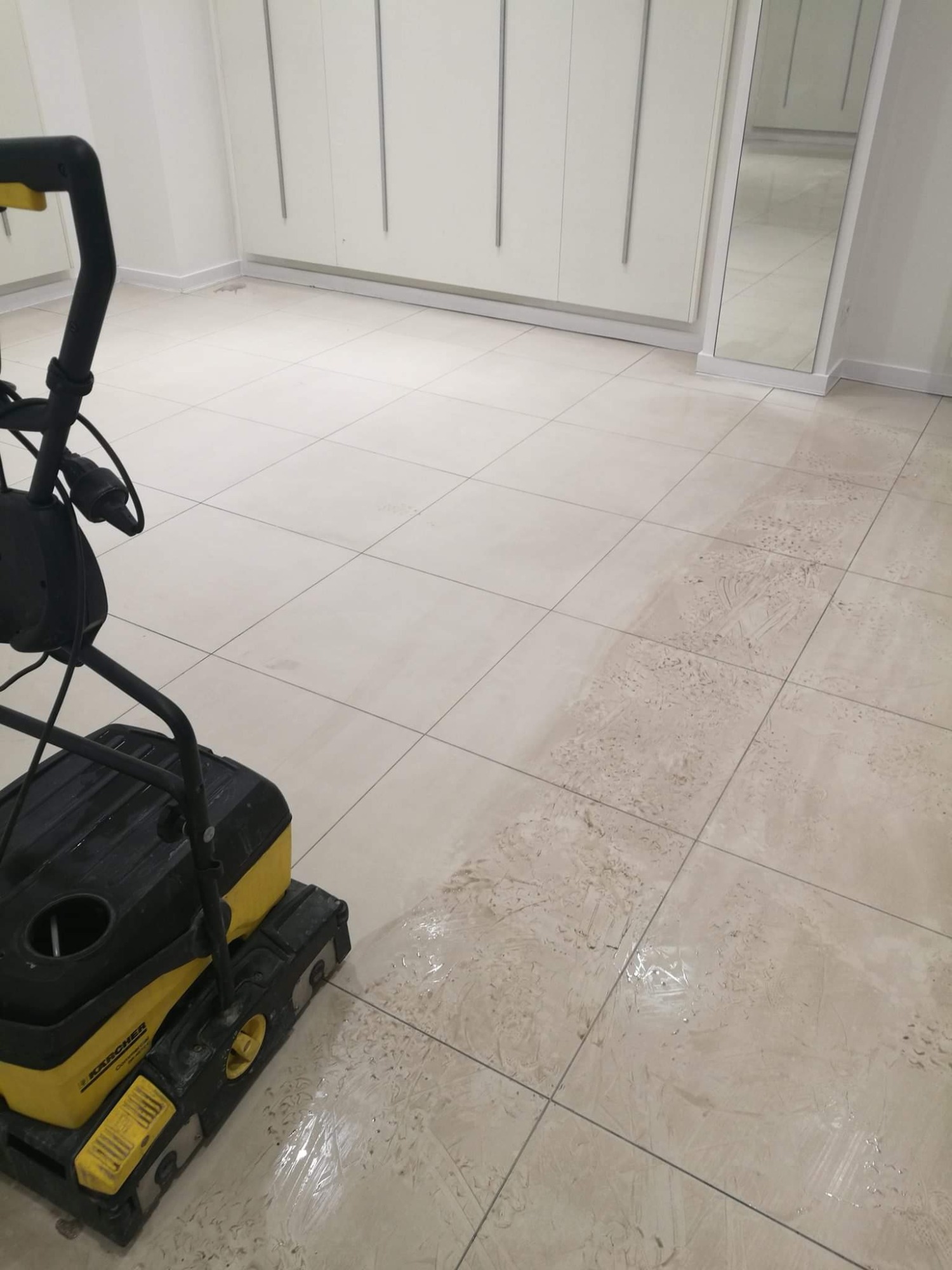 Profesjonalna maszyna Karcher do czyszczenia podłóg z płytek w trakcie pracy, widoczna różnica między czystą a brudną powierzchnią, tło stanowią białe szafki.