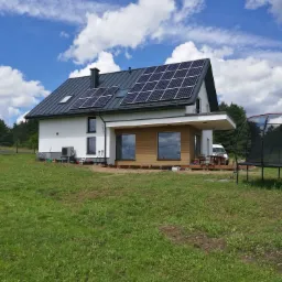 Nowoczesny dom jednorodzinny z panelami słonecznymi na ciemnoszarym dachu, białą elewacją i drewnianą dobud&oacute;wką, otoczony zielonym trawnikiem i błękitnym niebem z obłokami.