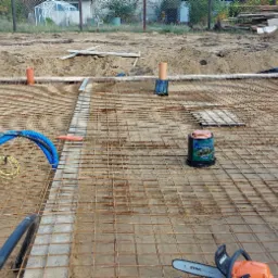 Zbrojenie fundamentu z siatki stalowej na przygotowanym podłożu, widoczne pionowe rury, niebieskie peszle, pojemnik z preparatem izolacyjnym Disprobit i piła spalinowa Stihl.