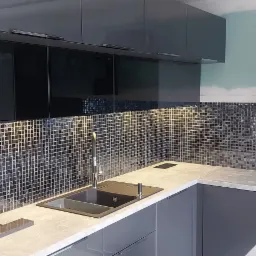 Nowoczesna kuchnia z szarymi szafkami, mozaikową ścianą nad blatem, czarnym zlewem i płytą gazową, okap Franke oraz niedokończone wykończenie ściany.