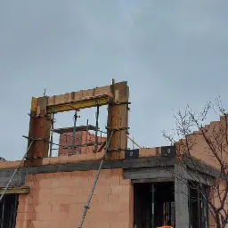 Wznoszenie ścian domu z pustaków ceramicznych, widoczne deskowanie i podpory szalunkowe, częściowo zapakowane pustaki na rusztowaniu wewnątrz budynku, biały samochód dostawczy zaparkowany obok.