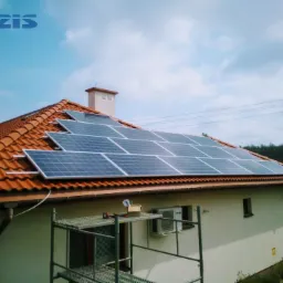 Panele słoneczne zamontowane na dachu domu z czerwoną dachówką, widoczne rusztowanie z narzędziami i klimatyzator na ścianie budynku.