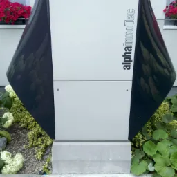 Zewnętrzna pompa ciepła Alpha InnoTec w obudowie z elementami grafitowymi, zamontowana na betonowej podstawie, otoczona roślinnością ogrodową, widok z poziomu gruntu.