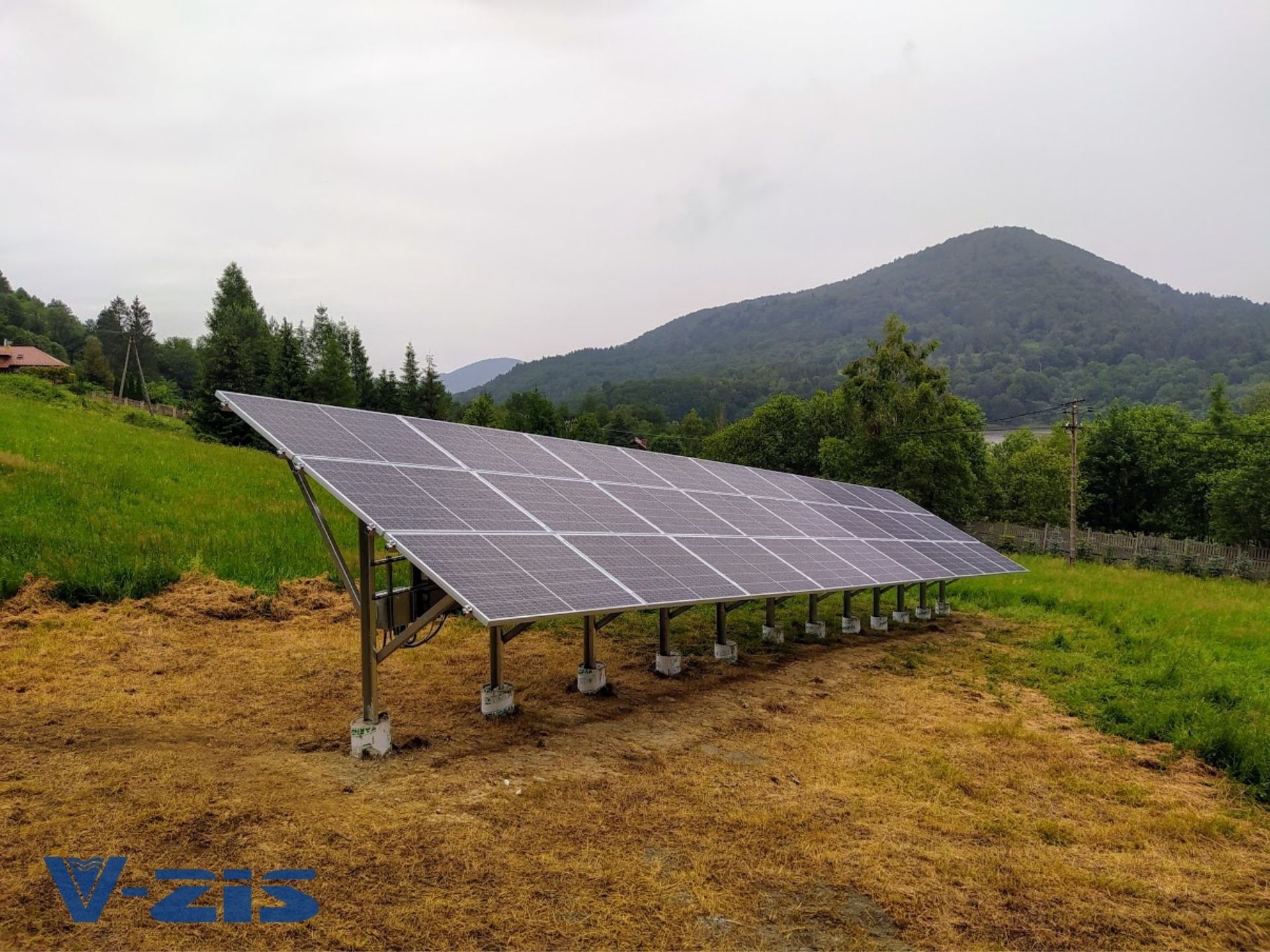 Rząd paneli słonecznych na metalowej konstrukcji, zamontowany na betonowych fundamentach, na tle zielonego wzgórza i zalesionych gór przy pochmurnej pogodzie.