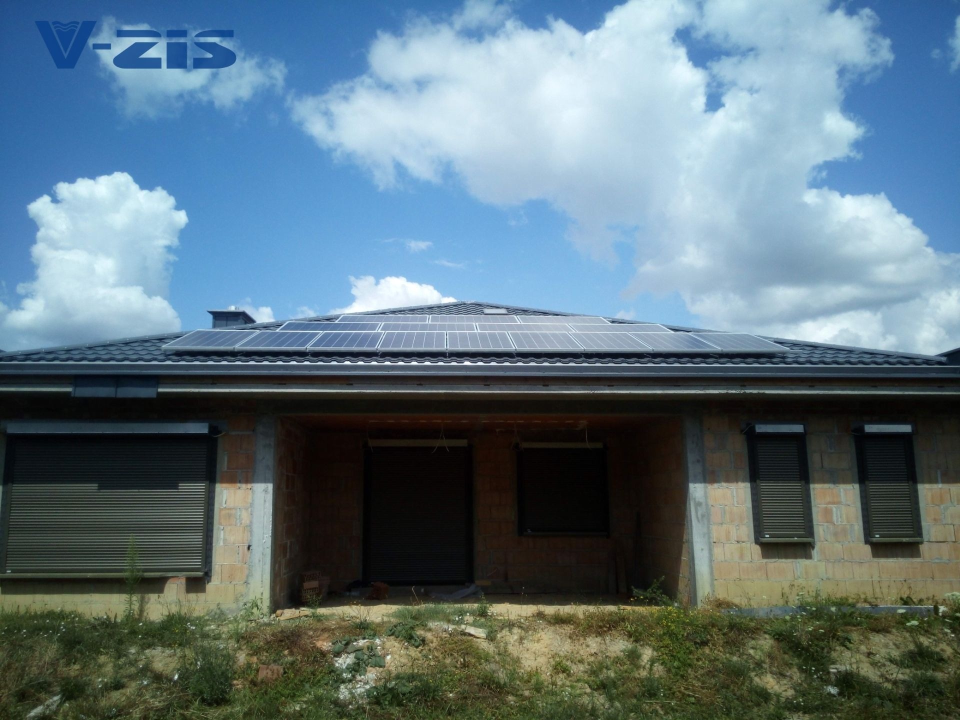 Niedokończony dom z cegły z szarymi roletami i panelami fotowoltaicznymi na dachu pod błękitnym niebem z chmurami, widoczne logo firmy V-zis.