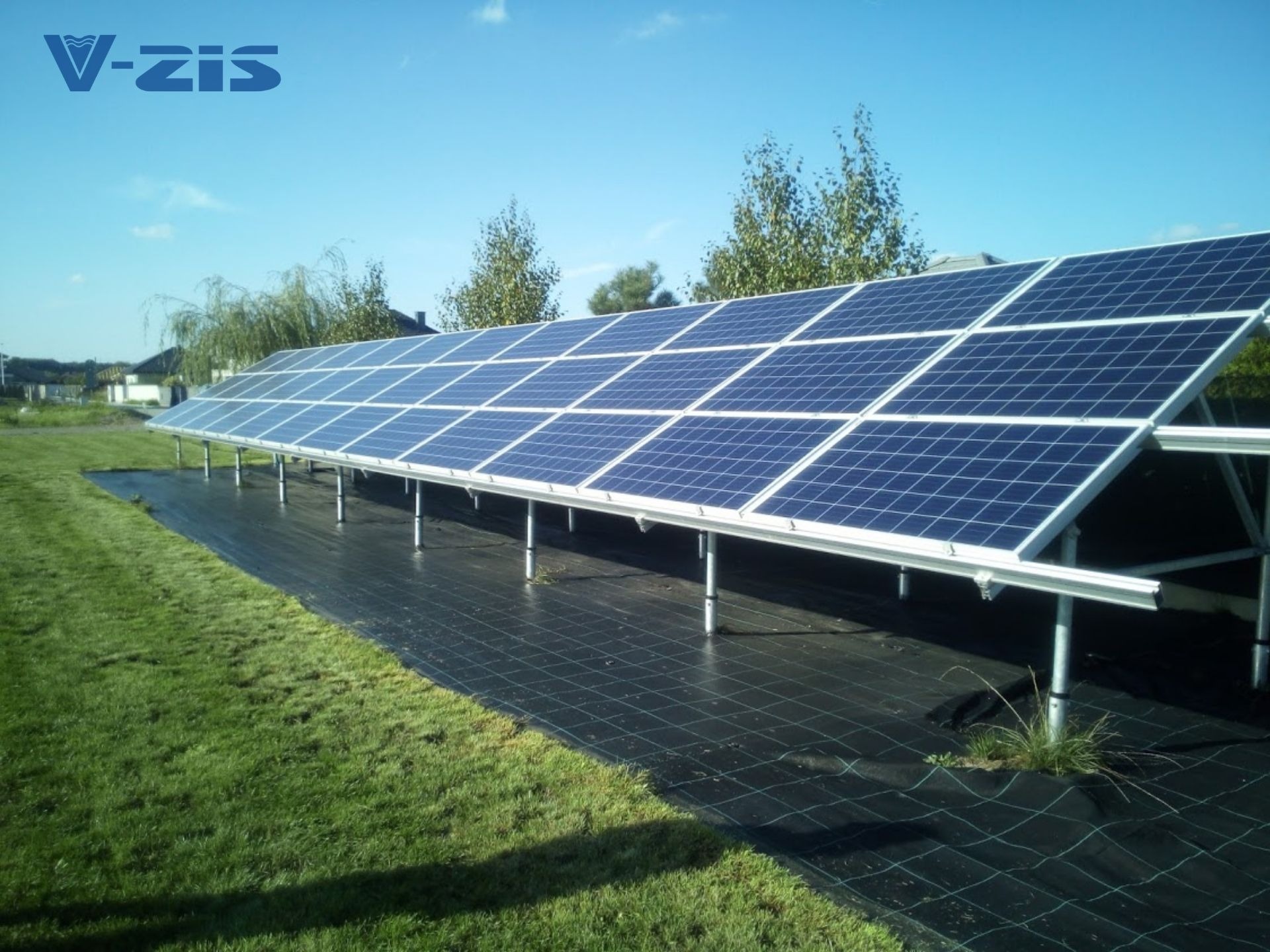 Rząd paneli słonecznych ustawionych na metalowych wspornikach nad czarną siatką na trawniku, widoczne logo firmy w górnym rogu.