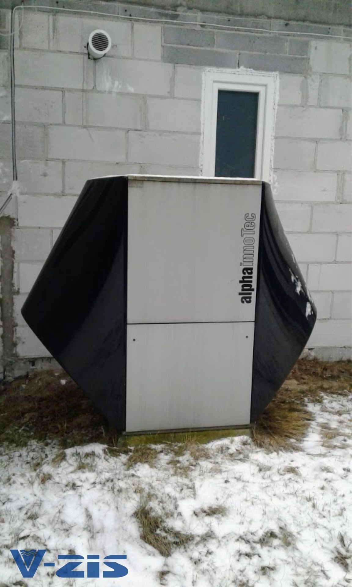 Kompaktowa instalacja grzewcza alpha innotec w czarno-białej obudowie, umieszczona na zewnątrz budynku z bloczków betonowych, w zimowej scenerii z resztkami śniegu.