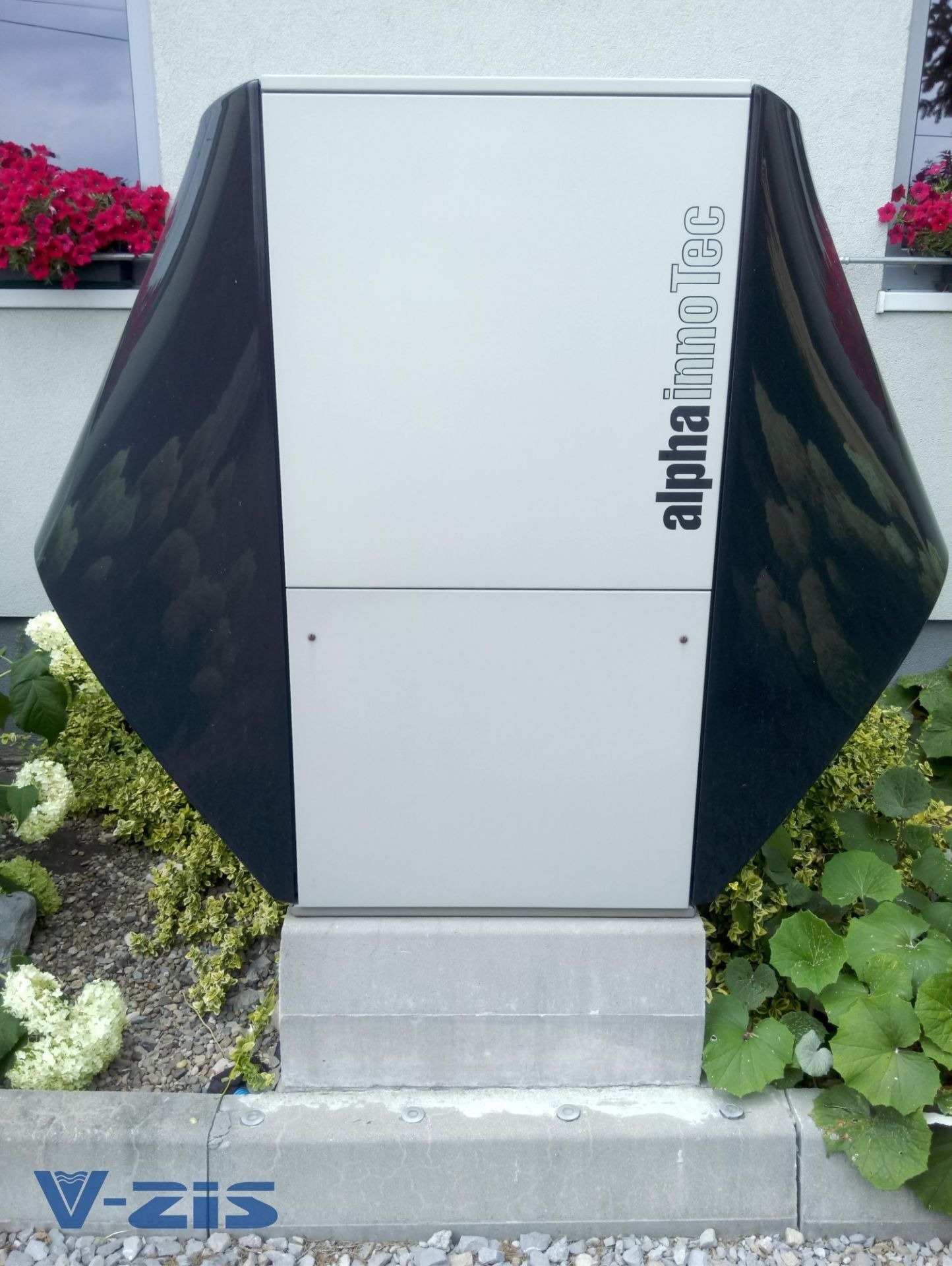 Zewnętrzna pompa ciepła Alpha InnoTec w obudowie z elementami grafitowymi, zamontowana na betonowej podstawie, otoczona roślinnością ogrodową, widok z poziomu gruntu.