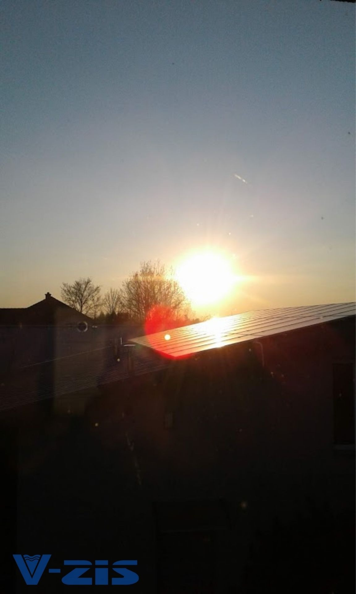 Panele słoneczne na dachu budynku oświetlone zachodzącym słońcem, widok z okna, logo firmy w dolnym rogu.