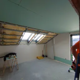 Poddasze w trakcie prac wykończeniowych: widoczne ocieplenie wełną mineralną, metalowy stelaż pod płyty, częściowo zamontowane płyty na suficie, okno dachowe, narzędzia i materiały na podłodze...