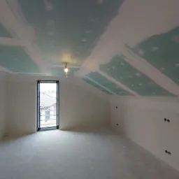 Poddasze po montażu płyt karton-gips i szpachlowaniu, widoczne łączenia płyt na suficie, surowy beton na podłodze i okno z widokiem na dach sąsiedniego budynku.