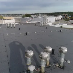 Widok z góry na płaski dach pokryty papą, z licznymi wywietrznikami turbinowymi i kominkami wentylacyjnymi. W tle zabudowania przemysłowe i drzewa.