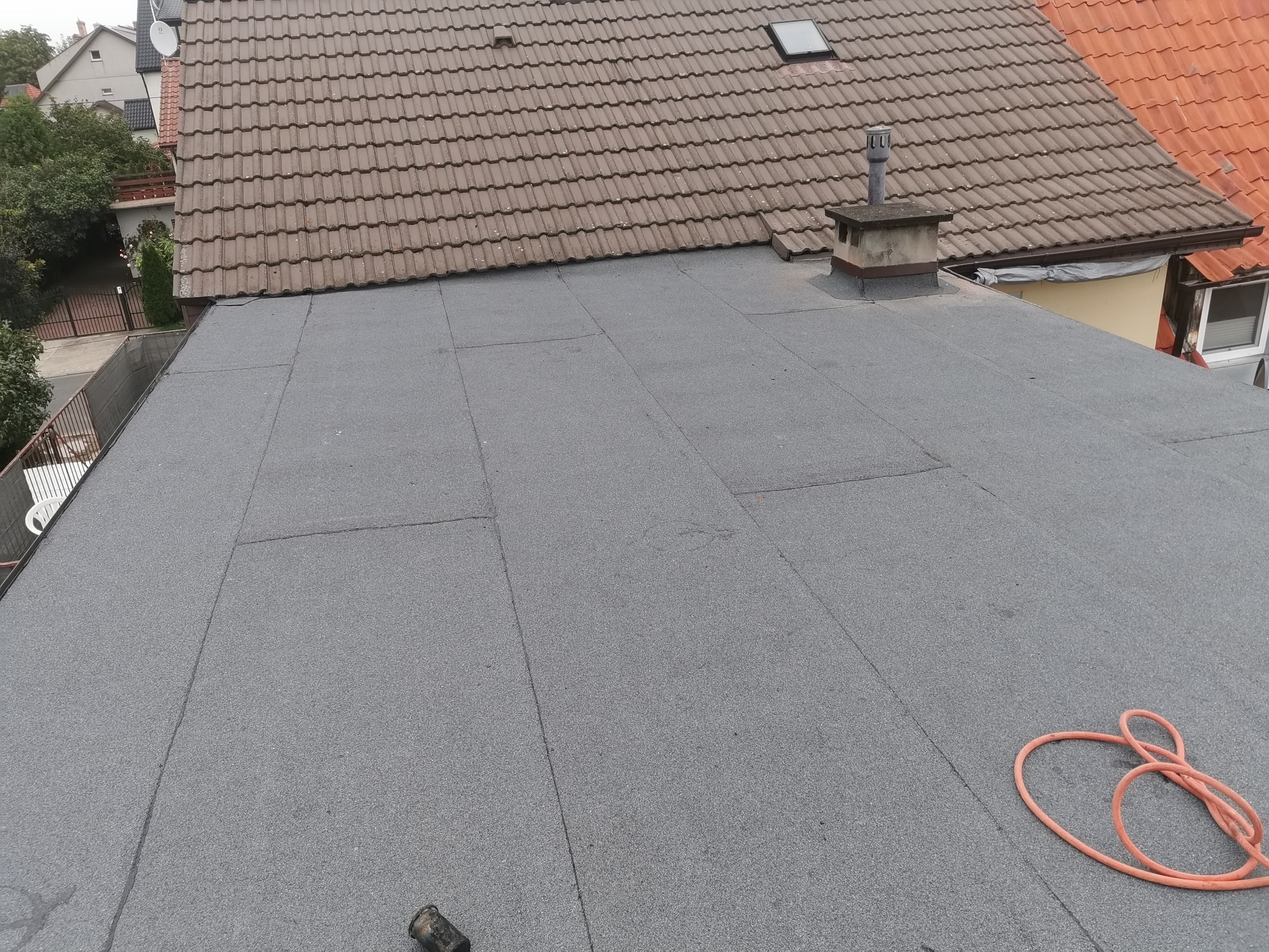 Płaski dach pokryty szarą papą bitumiczną, widoczny komin wentylacyjny i fragment dachu z dachówki ceramicznej w tle, obok leży zwinięty pomarańczowy wąż gumowy.