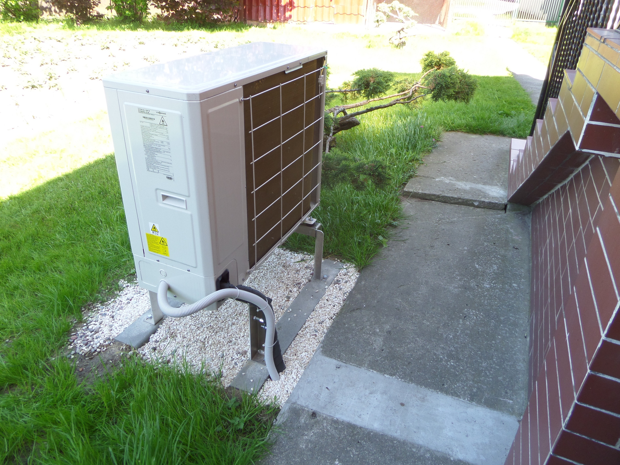 Zewnętrzna jednostka klimatyzacji ustawiona na betonowych podstawach, otoczona białym żwirem i trawą, z widocznymi przewodami i fragmentem schodów.