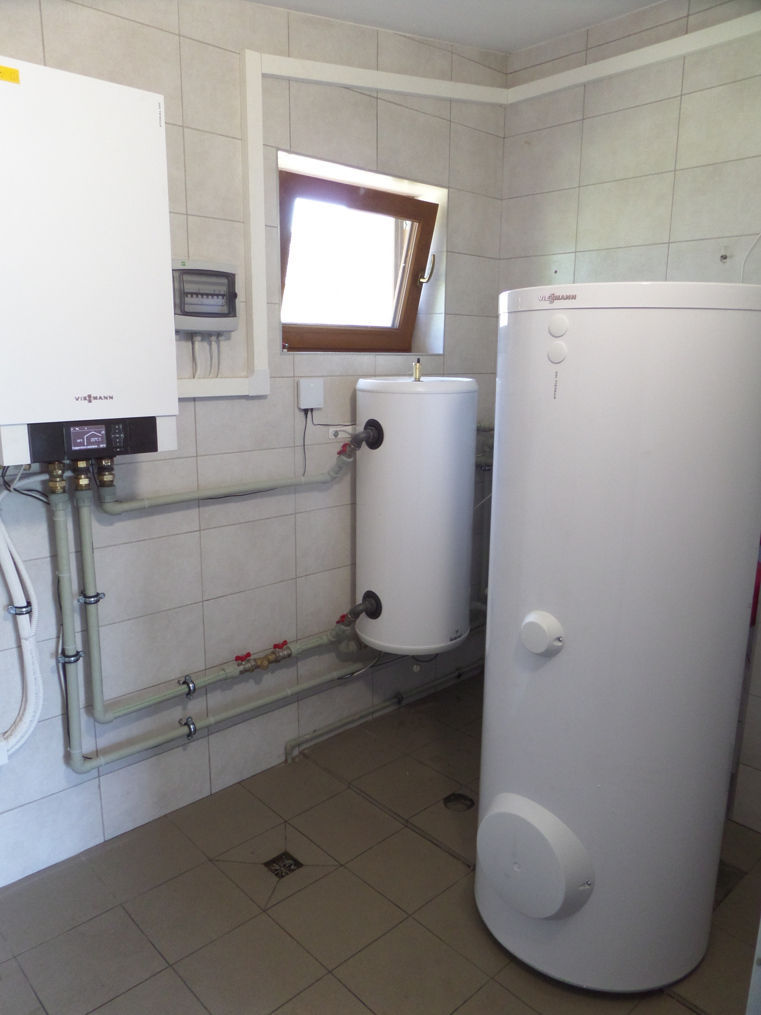 Biały piec gazowy Viessmann zamontowany na kafelkowanej ścianie w pomieszczeniu technicznym, z widocznymi rurami i dwoma zasobnikami ciepłej wody.