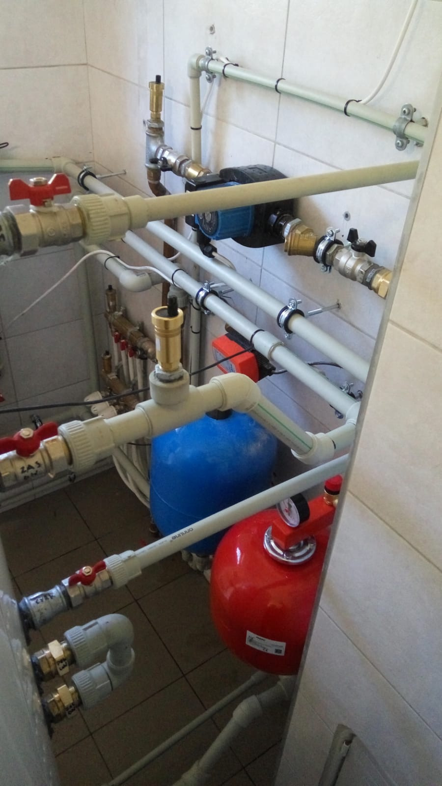 Instalacja z rur PVC, zaworów, pompy obiegowej i zbiorników wyrównawczych w kotłowni z białymi płytkami na ścianach.