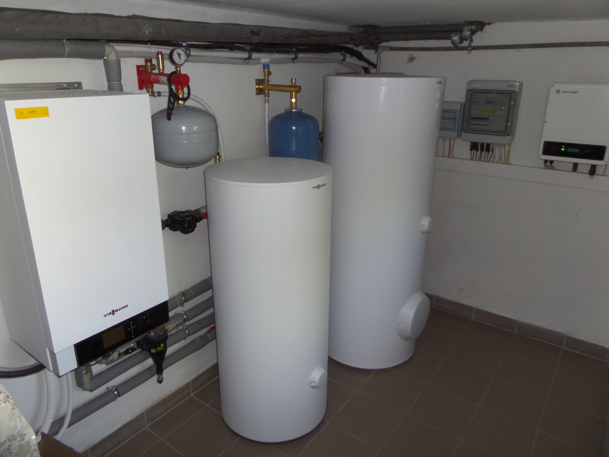 Kotłownia z białym kotłem grzewczym Viessmann, dwoma białymi zasobnikami ciepłej wody, szarą instalacją hydrauliczną i szarą skrzynką elektryczną na ścianie.