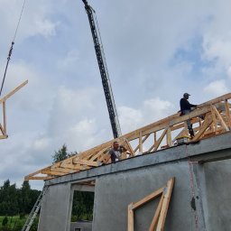 Budownictwo Systemowe Novum Sp. z o.o. - Montaż drewnianej więźby dachowej na domu z keramzytu przy użyciu dźwigu, widoczni dwaj pracownicy na dachu.