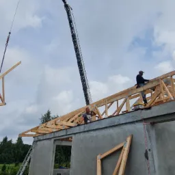 Montaż drewnianej więźby dachowej na domu z keramzytu przy użyciu dźwigu, widoczni dwaj pracownicy na dachu.