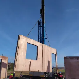 Montaż prefabrykowanej ściany z otworami okiennymi i drzwiowymi za pomocą dźwigu na placu budowy, widoczni pracownicy w kaskach i odzieży roboczej, słoneczny dzień.