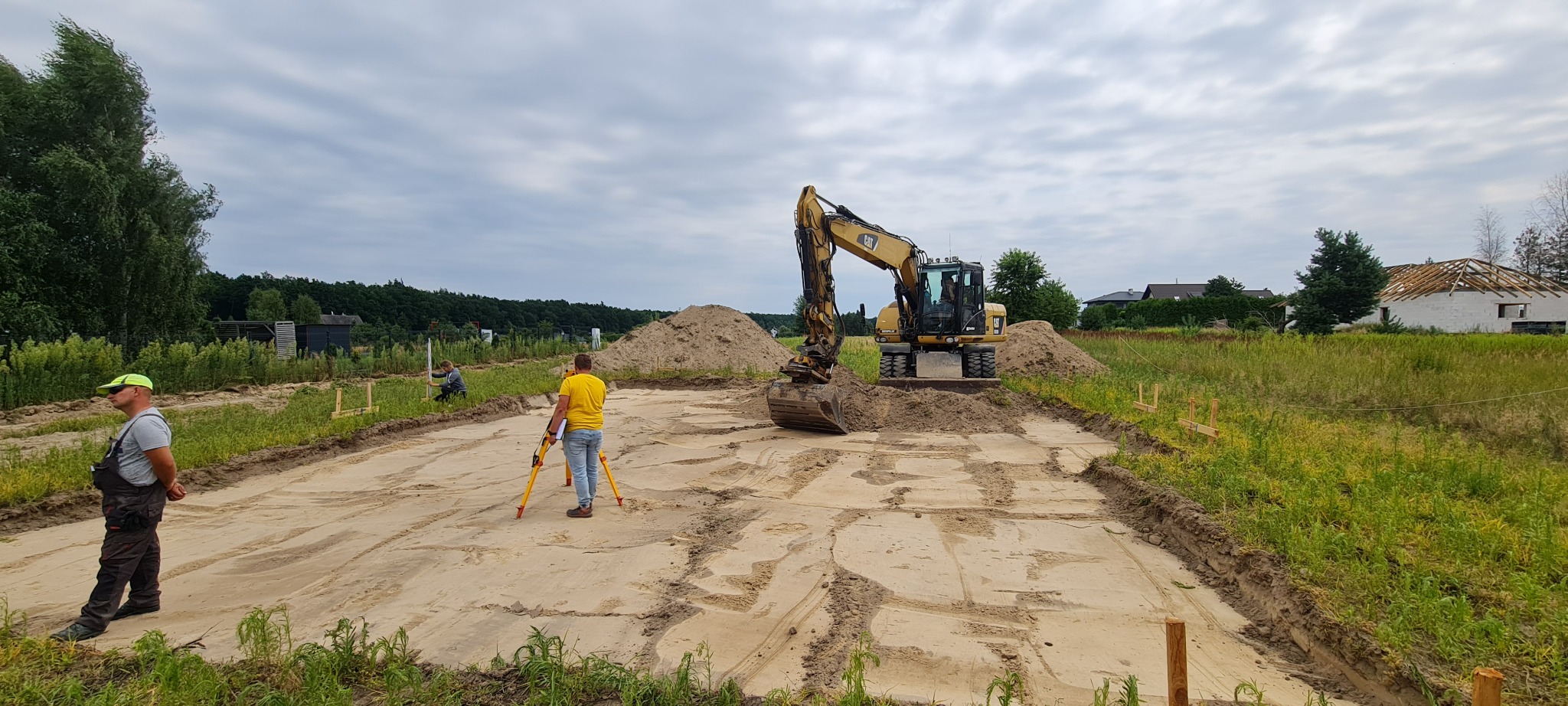 Przygotowanie terenu pod budowę domu: geodeta z niwelatorem, wykopany fundament i koparka na działce.