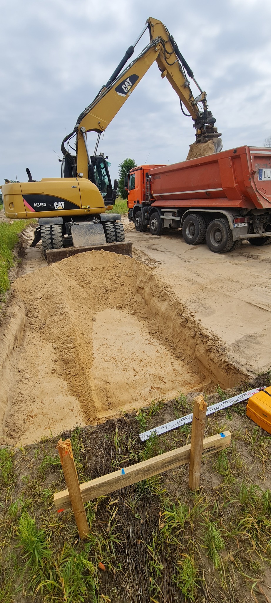 Żółta koparka CAT M316D nabiera piasek do pomarańczowej wywrotki na placu budowy; widoczne wykopane fundamenty i drewniane znaczniki.