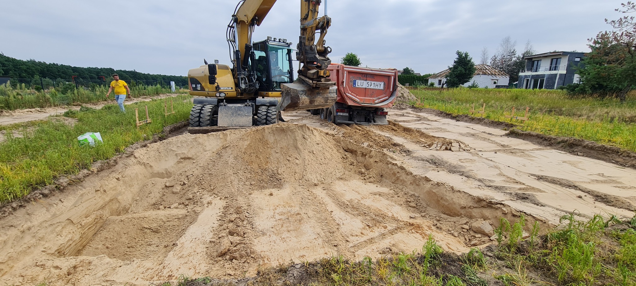 Żółta koparka nabiera piasek do wywrotki na placu budowy, w tle widoczny częściowo zadaszony dom w stanie surowym i mężczyzna w żółtej koszulce.