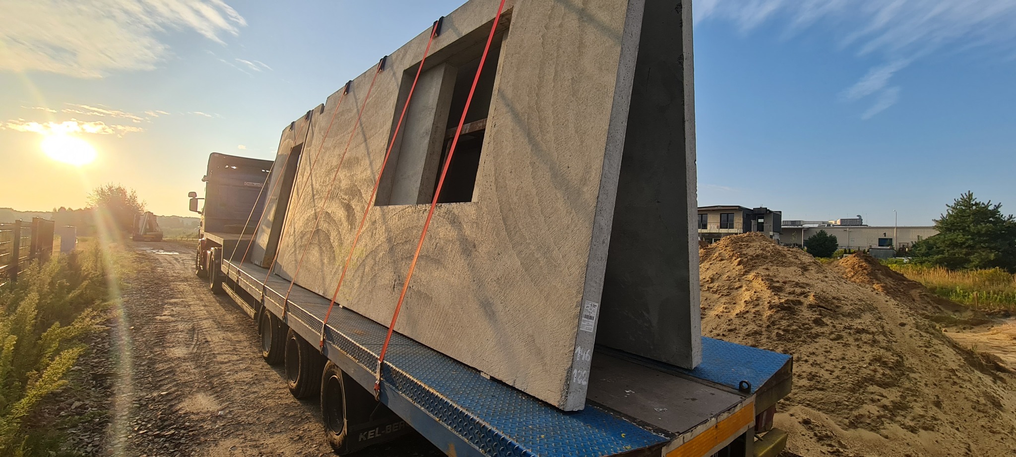 Transport prefabrykowanych elementów ściennych z otworami okiennymi na naczepie ciężarówki w Lublinie, z widocznym placem budowy w tle i zachodzącym słońcem.