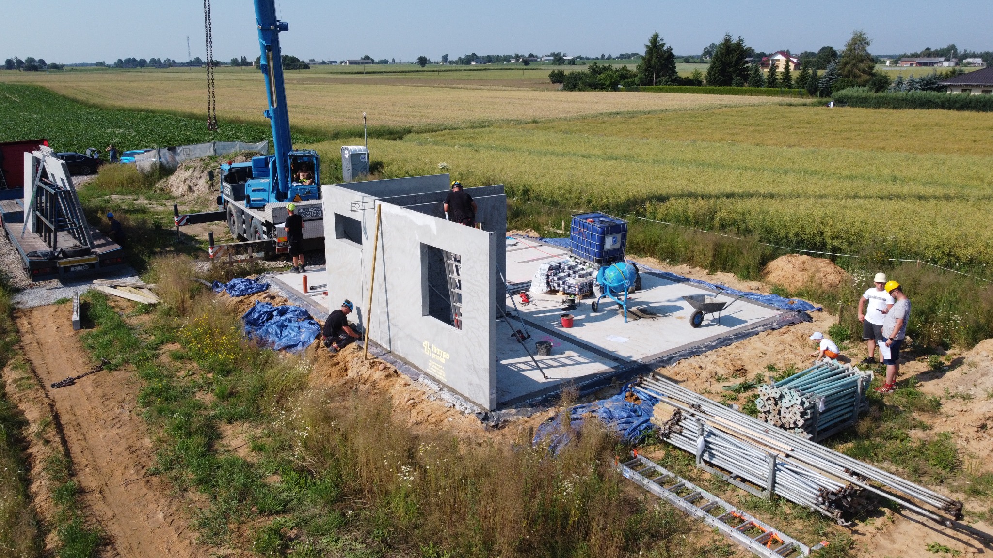 Montaż prefabrykowanych ścian domu na fundamencie, widoczny dźwig, mieszarka do betonu, pracownicy w kaskach, krajobraz polny w tle.