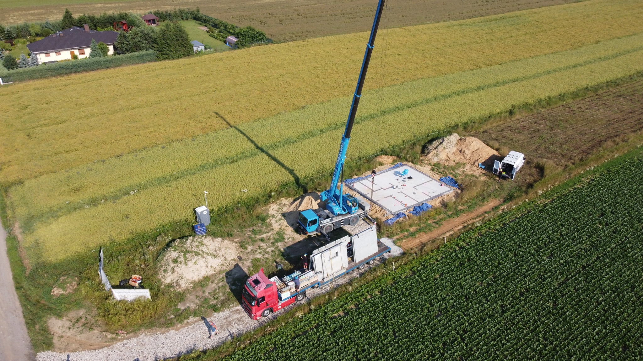 Widok z drona na transport i montaż domu modułowego na przygotowanym fundamencie w otoczeniu pól uprawnych, z użyciem dźwigu samochodowego.