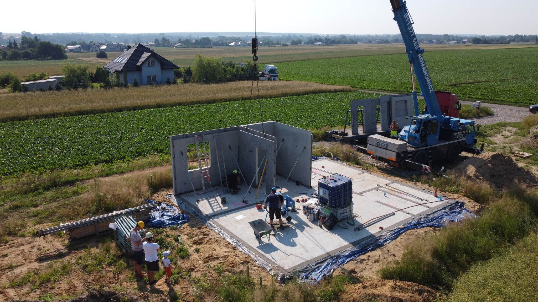 Montaż prefabrykowanych ścian domu na fundamencie, widok z góry na plac budowy z dźwigiem, grupa ludzi obserwuje postęp prac, w tle pola uprawne i domy.