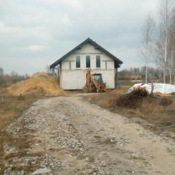 Jakub Kozakiewicz - Niedokończony dom jednorodzinny z trzema oknami na poddaszu, sterta piasku i koparka przed budynkiem, widok zza czarnego, metalowego ogrodzenia w pochmurny dzień.