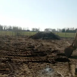 Żółta koparko-ładowarka na nierównym, brązowym terenie z widocznymi śladami opon, w tle hałda ziemi, siatka ogrodzeniowa i drzewa pod błękitnym niebem.