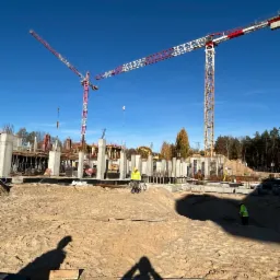 Budowa budynku żelbetowego w trakcie realizacji, widoczne szalunki, zbrojenia, betonowe słupy, dwóch pracowników w odblaskowych kamizelkach oraz dwa żurawie wieżowe na tle niebieskiego nieba i drzew.