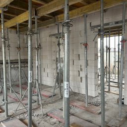 Jakub Kozakiewicz - Wnętrze budynku w trakcie budowy z białych bloczków, podparte metalowymi stemplami i żółtymi belkami stropowymi. Widoczne otwory okienne i drzwiowe.