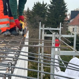 Jakub Kozakiewicz - Praca zbrojarska w Klukówku: zbrojarz w odzieży roboczej tnie pręt zbrojeniowy szlifierką kątową na tle konstrukcji zbrojeniowej i zieleni.