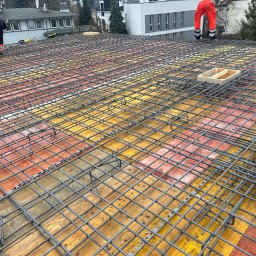 Jakub Kozakiewicz - Siatka zbrojeniowa na deskowaniu, przygotowana do zalania betonem. W tle pracownicy w odzieży roboczej. Widok z góry, szeroki kadr.