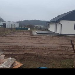 Jakub Kozakiewicz - Nowo wybudowany dom jednorodzinny z ciemnym dachem i jasną elewacją, widoczny z boku, z przygotowanym terenem wokół, częściowo utwardzonym kostką, w pochmurny dzień.