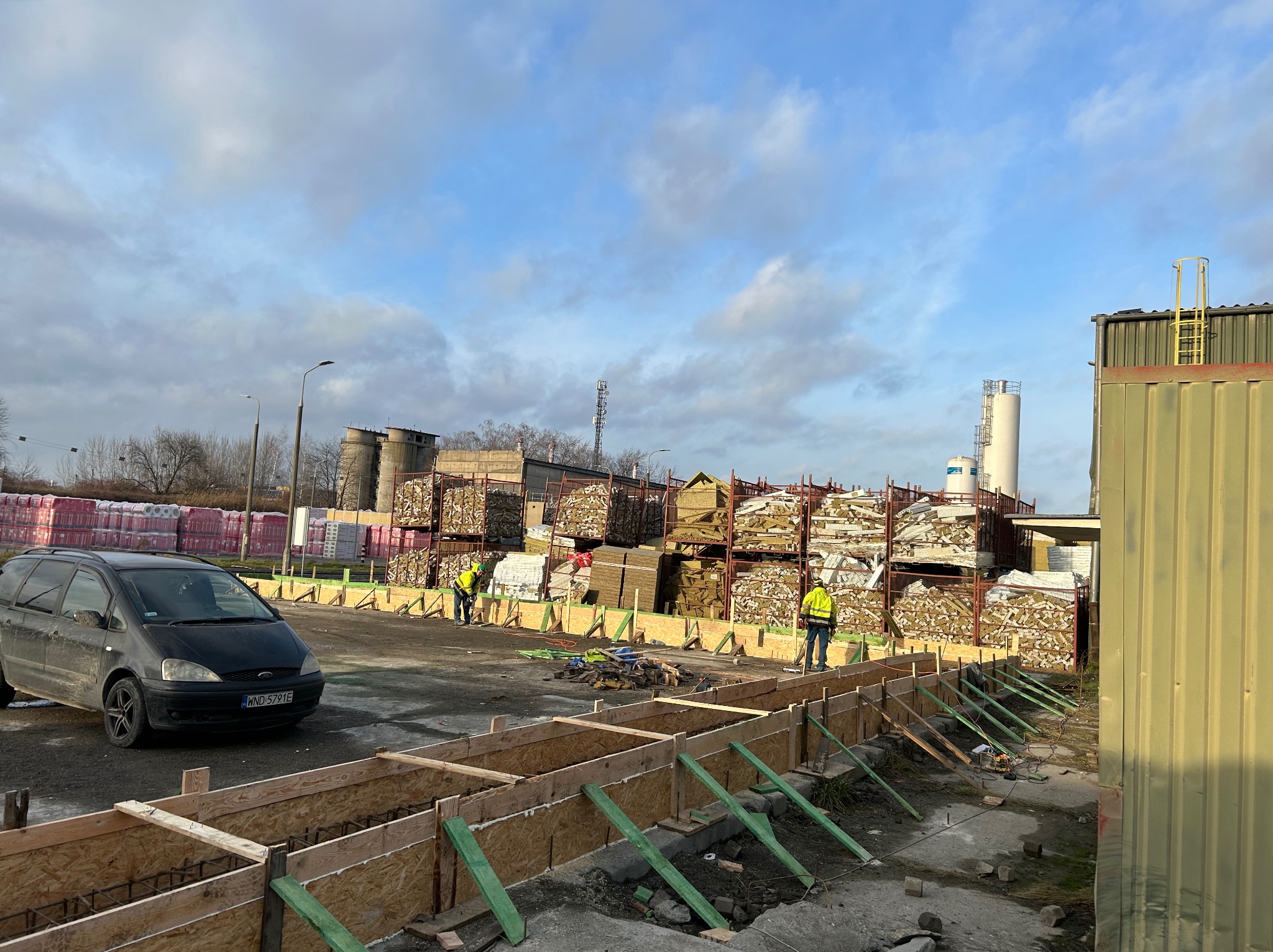 Przygotowanie szalunku pod wylewkę fundamentów z widocznym zbrojeniem, pracownicy w odblaskowych kamizelkach i sterty materiałów budowlanych w tle.